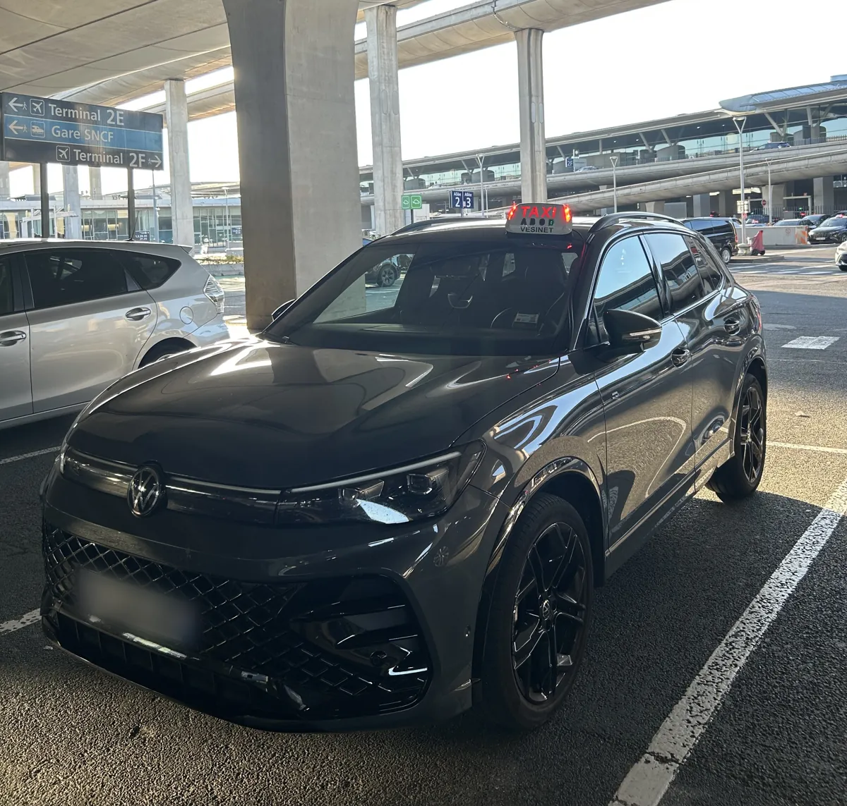 Le taxi de Samy garé au terminal 2 de Roissy-Charles de Gaulle, panneaux Terminal 2E et 2F au fond